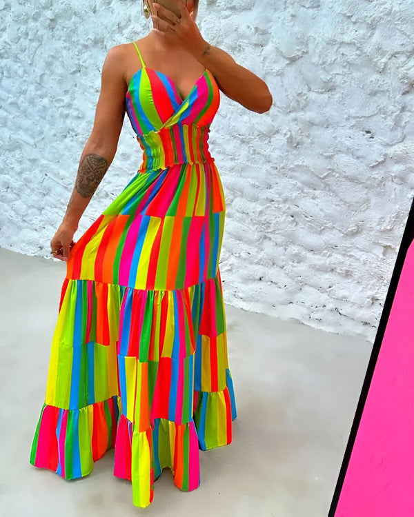Une femme porte une robe longue et colorée. La robe est à fines bretelles et a un décolleté en V. Elle est cintrée à la taille et évasée sur le bas. La robe est multicolore, avec des rayures verticales de différentes couleurs. La femme est debout devant un mur blanc.