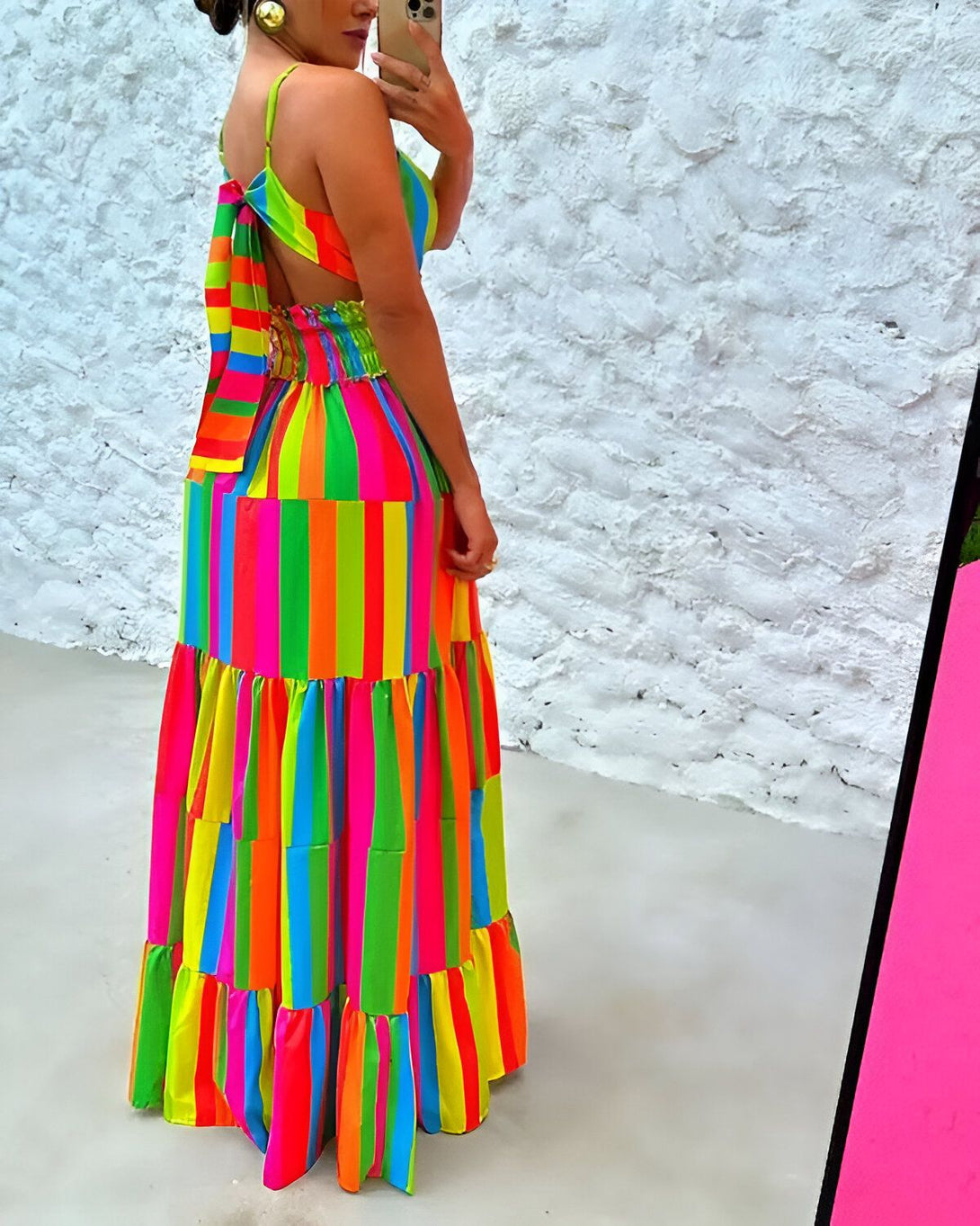 Une femme porte une robe longue à fines bretelles et à rayures verticales multicolores. La robe est ouverte dans le dos et nouée au niveau de la taille. La femme est vue de dos et porte des boucles d'oreilles rondes et un bracelet. Elle tient un téléphone portable dans sa main droite et se prend en photo dans un miroir.