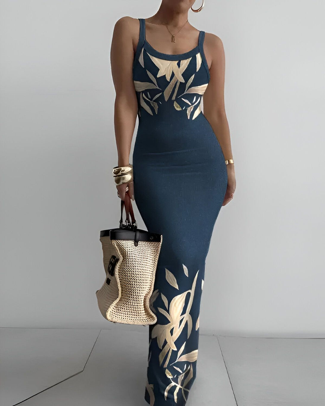 Une femme porte une robe longue bleu marine près du corps à fines bretelles. La robe est imprimée de feuilles tropicales dorées. Elle porte également un sac à main en paille beige et des bijoux dorés.