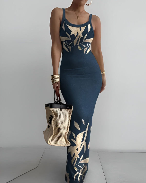 Une femme porte une robe longue bleu marine près du corps à fines bretelles. La robe est imprimée de feuilles tropicales dorées. Elle porte également un sac à main en paille beige et des bijoux dorés.