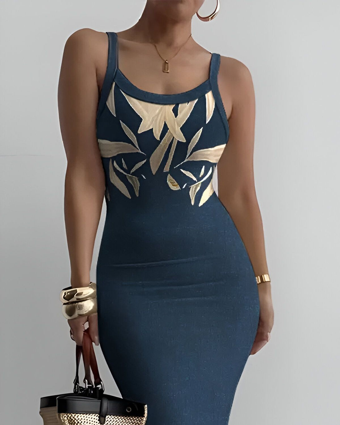 Une femme porte une robe moulante bleu marine à bretelles avec un motif de feuilles tropicales dorées. Elle porte également des bracelets dorés et tient un sac à main marron.