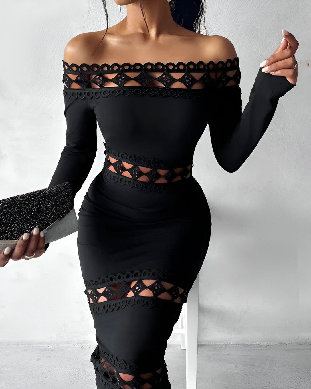 Une femme porte une robe noire moulante avec des découpes au niveau de la poitrine et des hanches. Elle a les cheveux longs et noirs et porte un maquillage glamour. Elle est debout dans une pièce blanche et tient un sac à main noir.