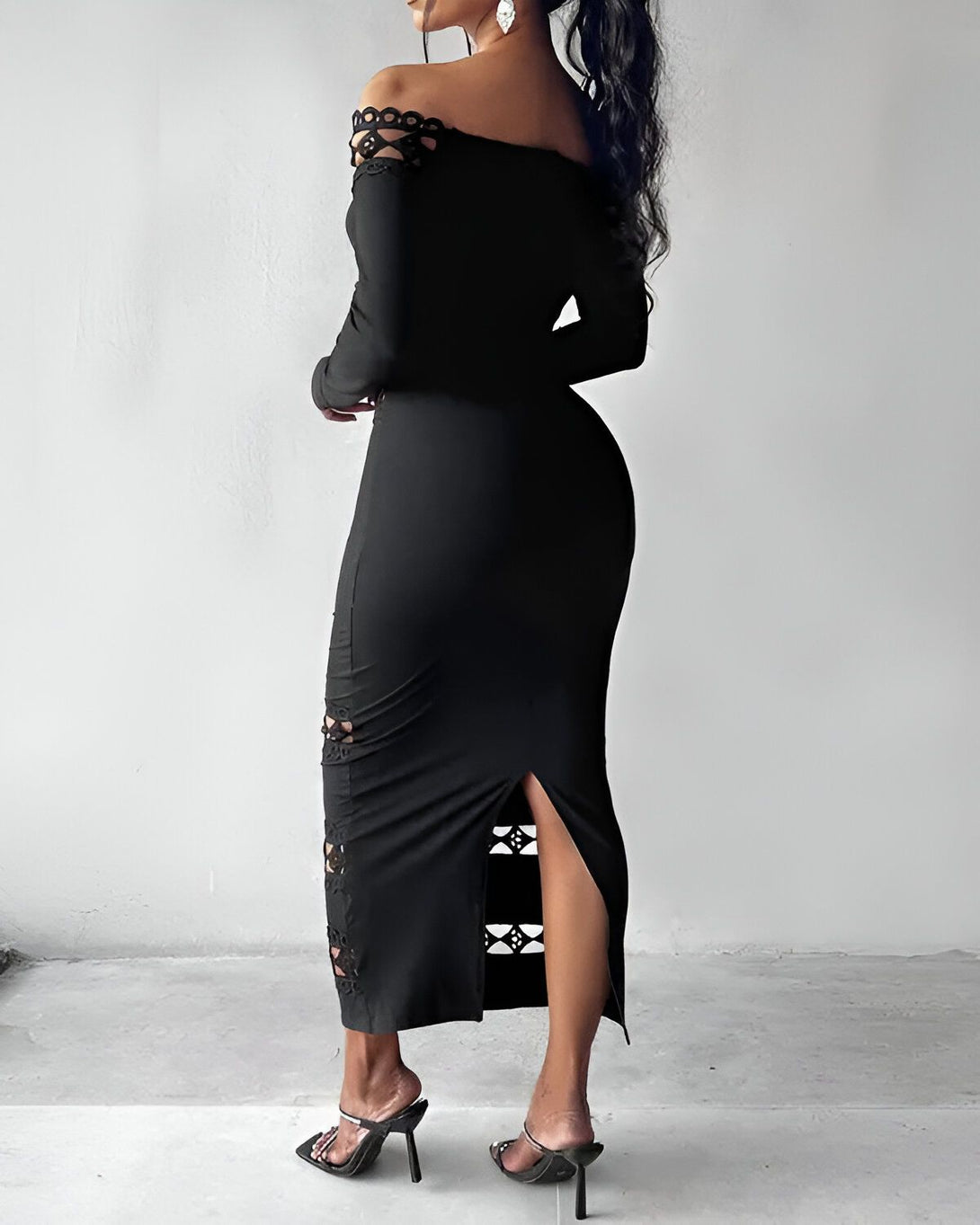 Une femme porte une robe noire moulante à épaules dénudées. La robe est fendue sur le côté et possède des détails ajourés. La femme est vue de dos et porte des talons noirs.