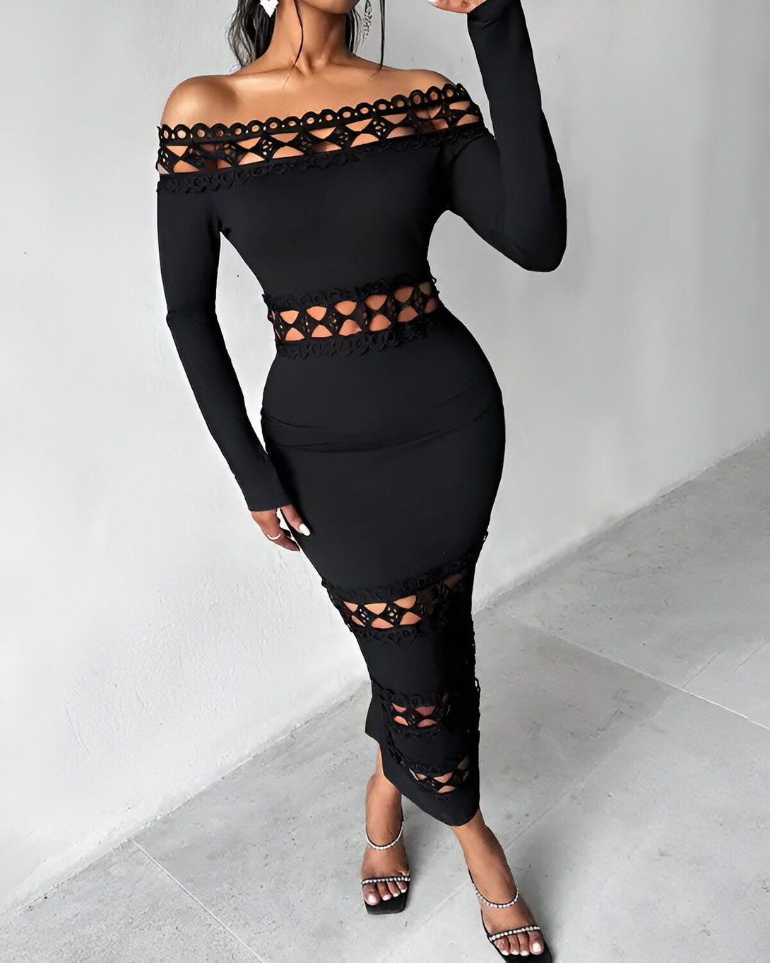 Une femme porte une robe noire moulante. La robe a un décolleté en cœur et des manches longues. Elle est ouverte au niveau de la taille et des hanches. La femme porte des escarpins noirs et des boucles d'oreilles en diamant.