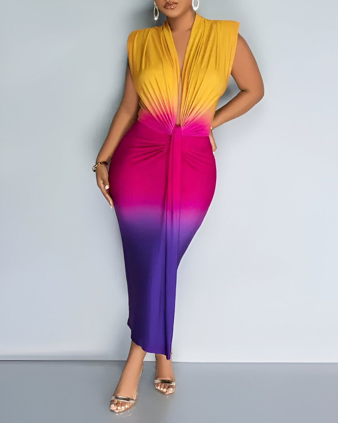 Une femme porte une robe moulante jaune, rose et violet. La robe est nouée sur le devant et a une fente sur la jambe droite. La femme porte des sandales à talons argentées et des boucles d'oreilles.