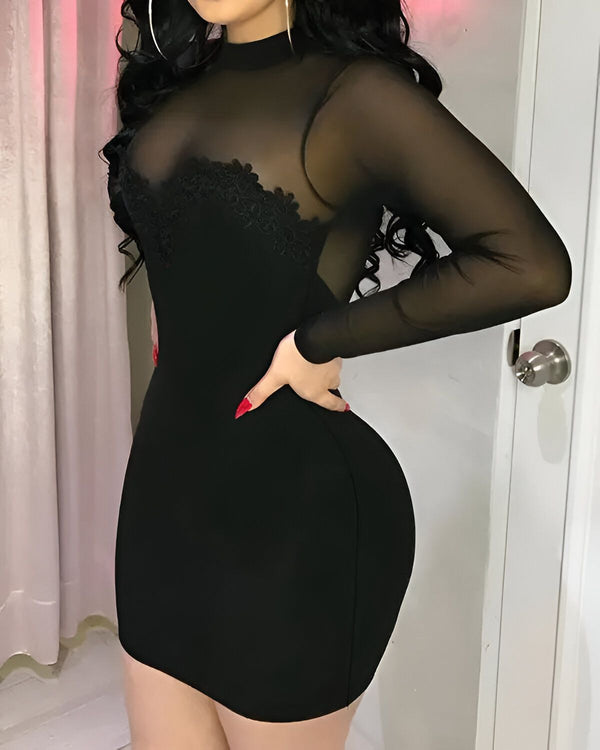 Une femme porte une robe moulante noire avec un col montant et des manches longues. La robe est transparente au niveau du buste et laisse entrevoir un soutien-gorge noir. La femme a les cheveux longs et noirs et porte des talons noirs. Elle est debout dans une pièce avec un mur rose et une porte blanche.