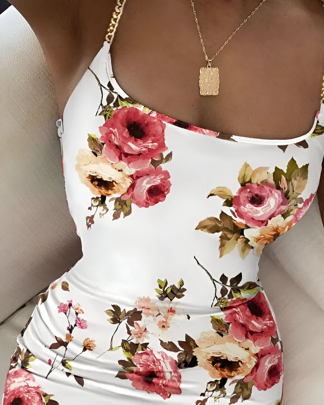 Une femme porte une robe blanche moulante à fleurs roses et rouges. Elle porte également un collier en or avec un pendentif carré.