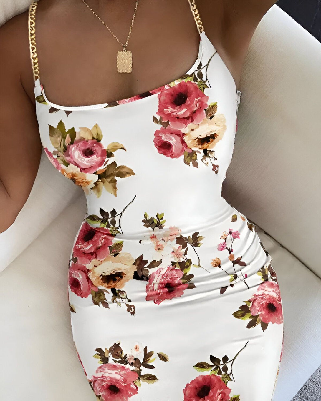 Une femme porte une robe blanche moulante à fleurs roses et rouges. Elle a un collier avec un pendentif doré. Ses cheveux sont noirs et elle a les lèvres rouges. Elle est assise sur un canapé blanc.