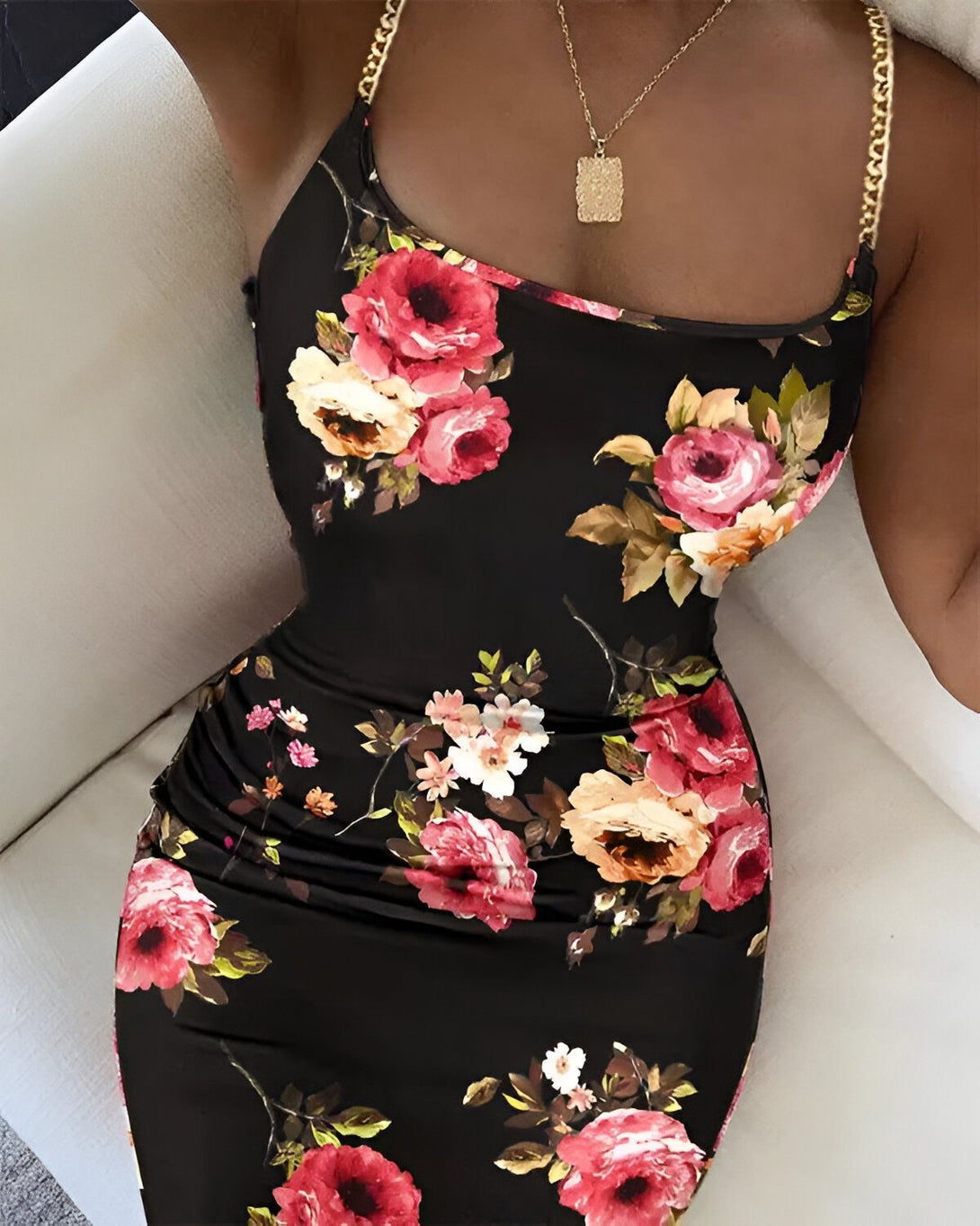 Une femme porte une robe noire moulante à fleurs. La robe a un décolleté en cœur et des bretelles fines. La femme est assise sur un canapé blanc.