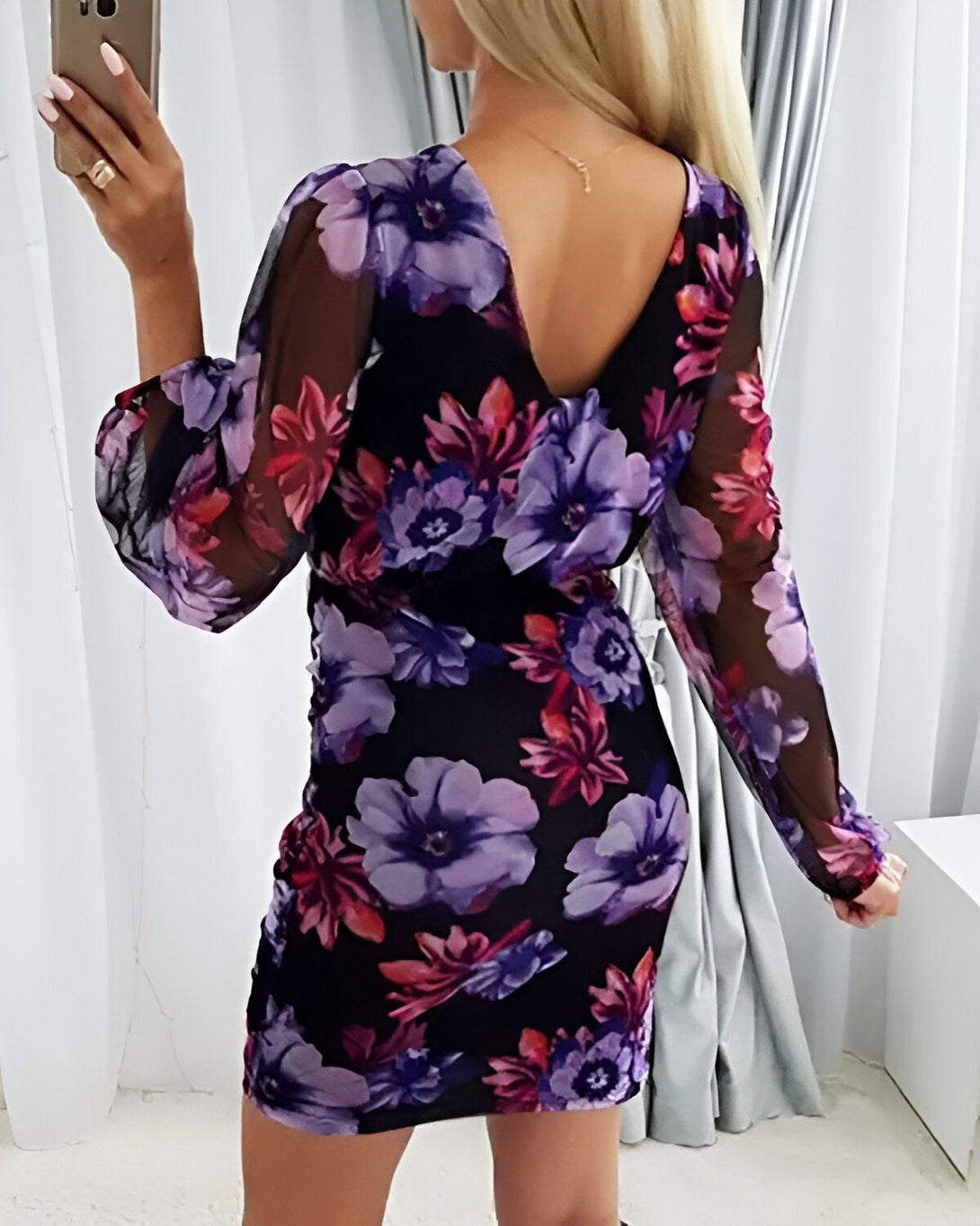 Une femme porte une robe moulante noire à fleurs mauves et roses. La robe est décolletée dans le dos et possède des manches longues bouffantes.