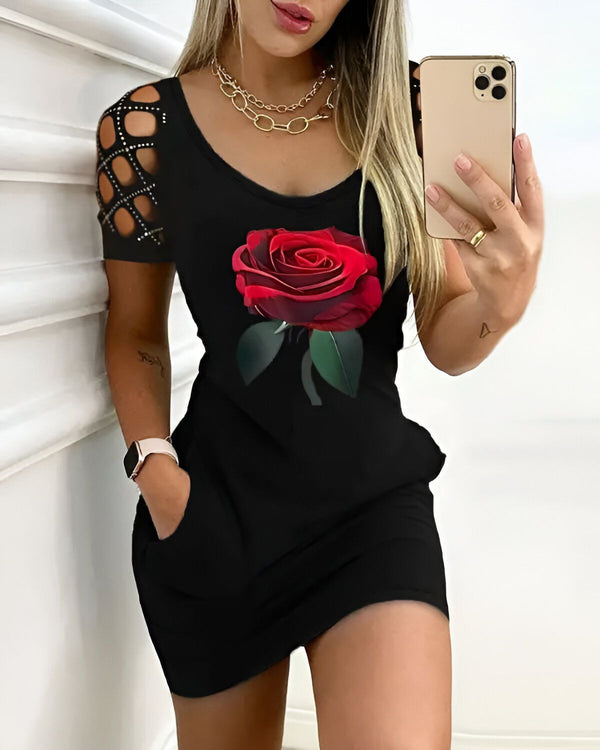 Une jeune femme porte une robe noire moulante avec un imprimé de rose rouge. La robe a des manches courtes et un col rond. La femme a les cheveux longs et blonds et elle est maquillée. Elle porte un collier et un bracelet. Elle est debout devant un mur blanc et elle tient un téléphone portable dans sa main droite.