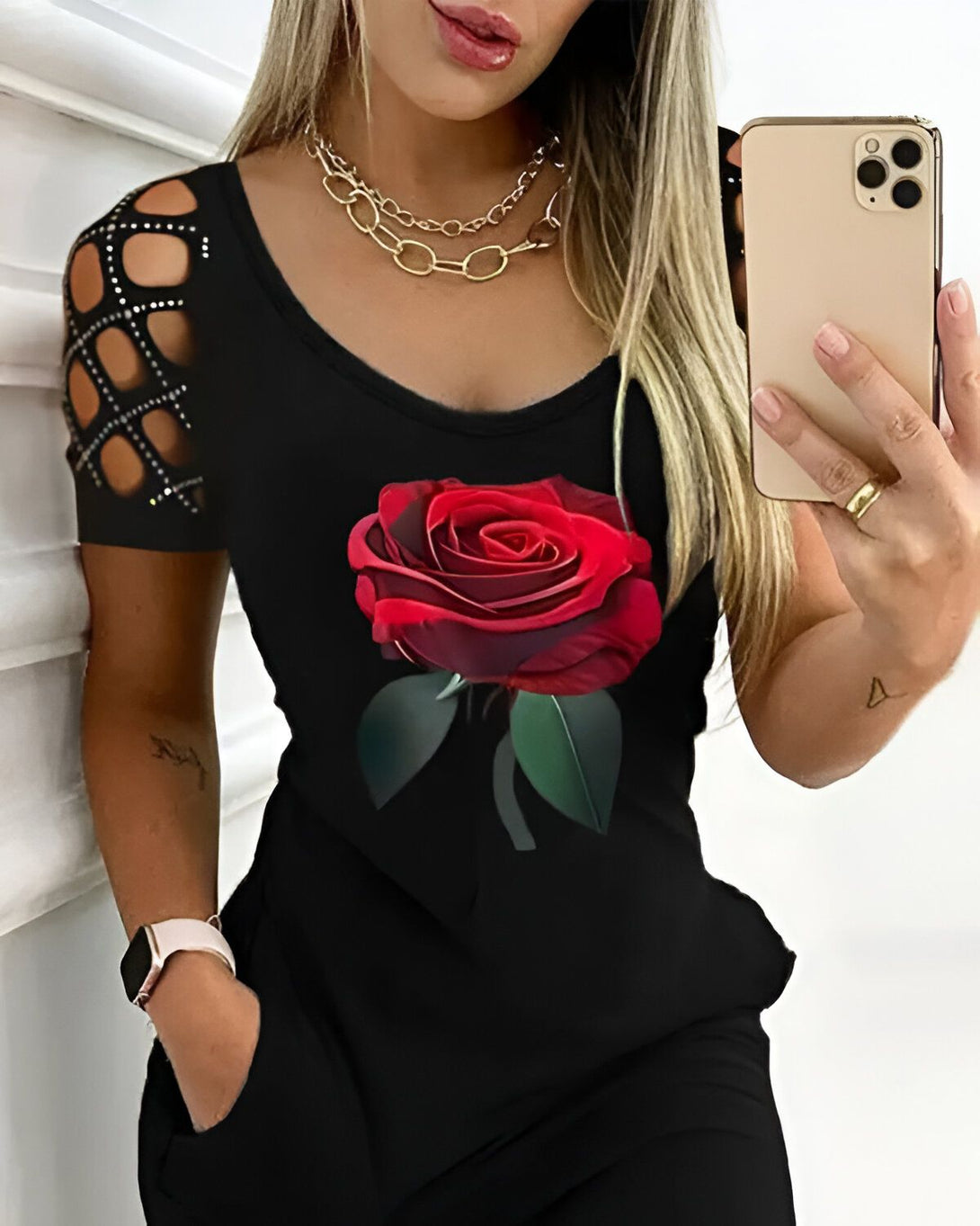 Une femme porte une robe noire avec une image d'une rose rouge imprimée sur le devant. La robe a des manches courtes et un col rond. La femme a les cheveux longs et noirs et elle porte un collier. Elle tient un téléphone portable dans sa main droite.