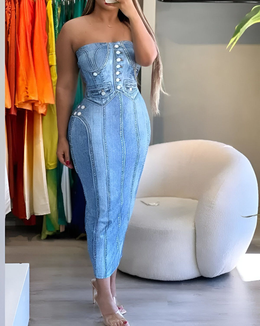 Une femme porte une robe en jean bleu clair. La robe est sans bretelles et a une coupe ajustée. Elle est boutonnée sur le devant et a des poches sur les côtés. La femme a les cheveux longs et bruns et porte des talons hauts transparents. Elle se tient debout dans un magasin de vêtements et il y a des vêtements colorés sur des portants derrière elle.