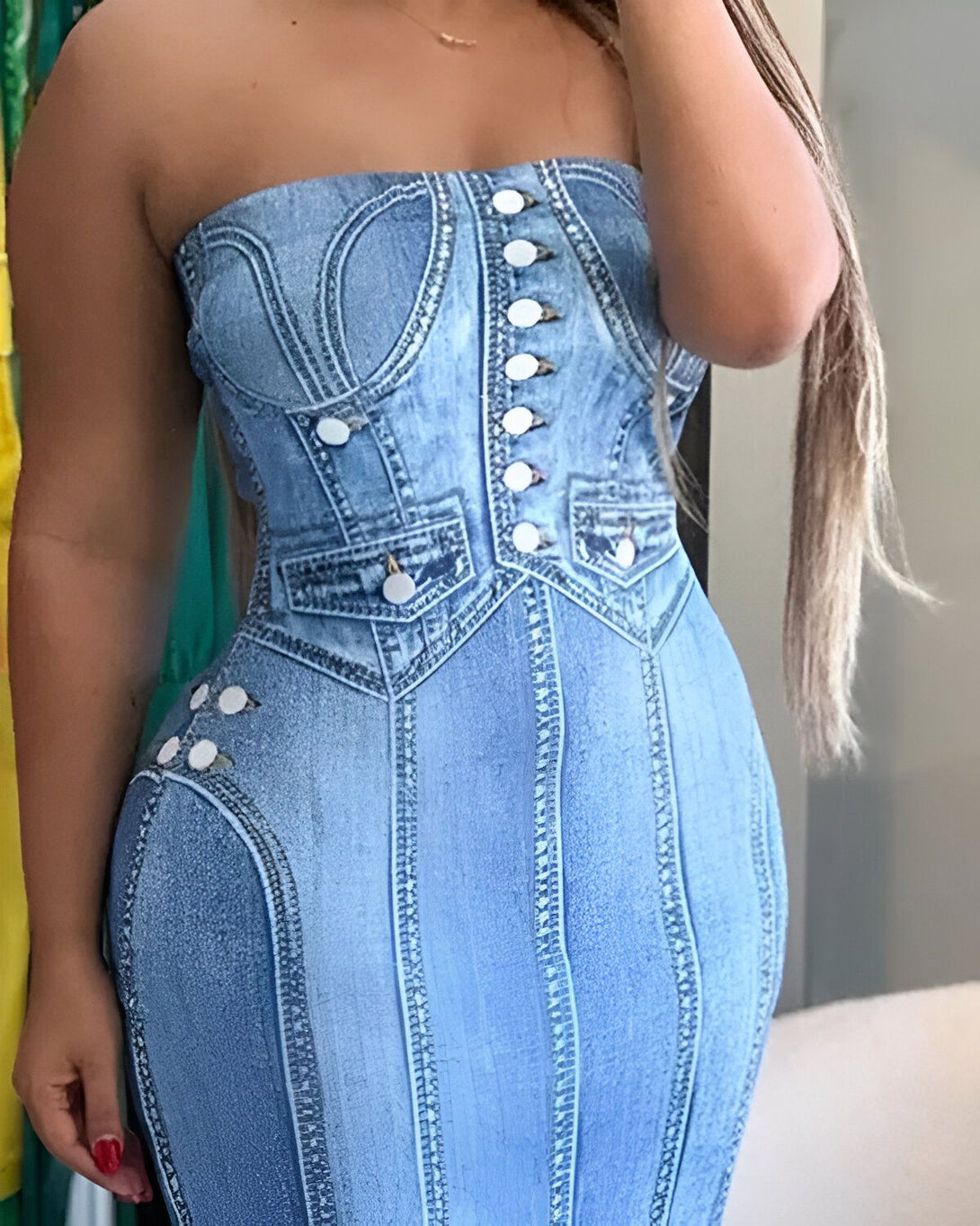 Une femme porte une robe en jean strapless. La robe est ajustée et a des boutons sur le devant. La femme a les cheveux longs et noirs et porte du maquillage. Elle est debout dans un magasin et sourit à la caméra.