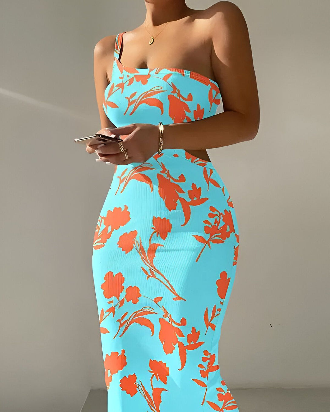 Une femme porte une robe longue moulante à une épaule de couleur bleu clair avec des motifs de fleurs oranges. Elle a un téléphone portable à la main.