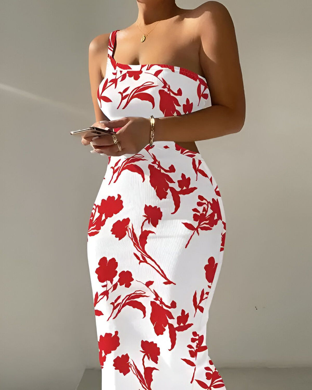 Une femme porte une robe longue blanche à motifs de fleurs rouges. Elle est debout, de profil, et tient un téléphone portable dans sa main droite.