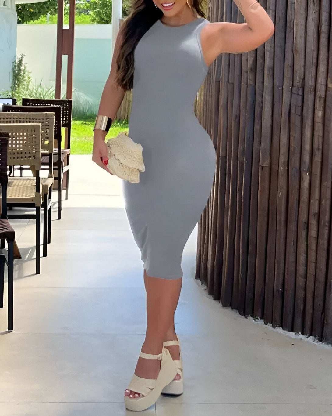 Une femme porte une robe moulante gris clair. Elle est debout dans un couloir avec des chaises et des tables en arrière-plan. Elle porte des sandales à talons compensés couleur chair et tient un sac à main beige.