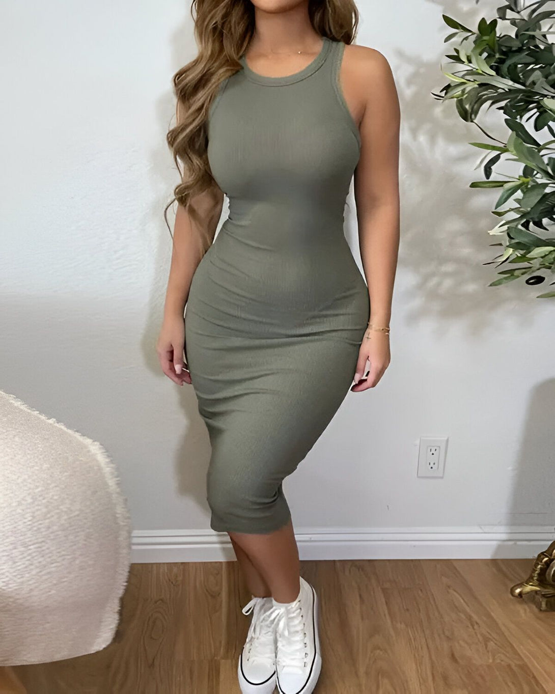 Une femme porte une robe vert olive moulante et des baskets blanches. Elle est debout dans un salon, il y a un arbre en pot et un tapis beige.
