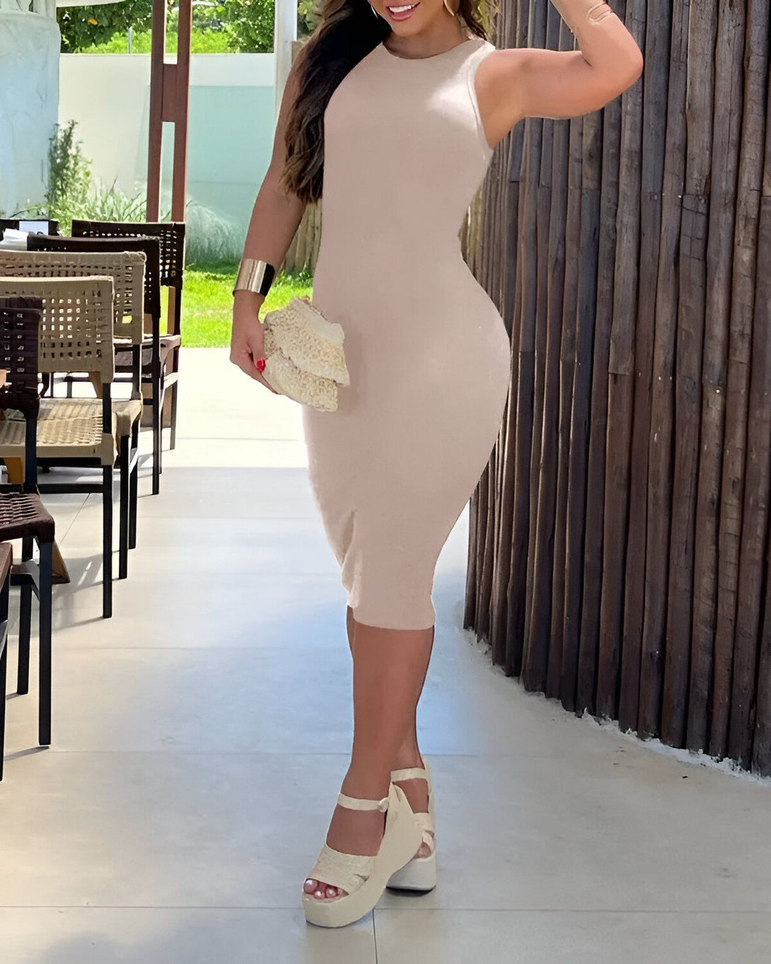 Une femme porte une robe beige moulante et des sandales compensées beiges. Elle a un sac à main en osier à la main et des bracelets aux poignets.