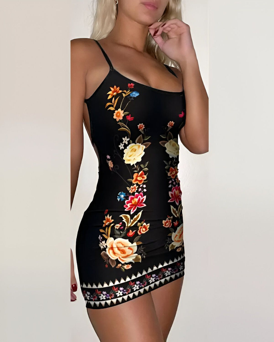Une jeune femme blonde porte une robe moulante noire à motifs de fleurs colorées. La robe est courte et met en valeur ses jambes. Elle porte des talons hauts noirs et un collier ras du cou. Ses cheveux sont lâchés et elle a un maquillage léger.