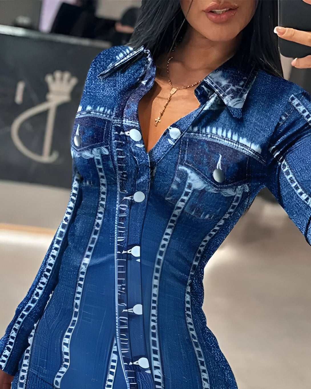 Une femme porte une robe chemise en jean bleu. La robe est cintrée et a un col montant. Elle est ouverte sur le devant et on peut voir son décolleté. La femme a les cheveux longs et noirs et elle porte un collier avec une croix. Elle est maquillée et a les lèvres rouges.