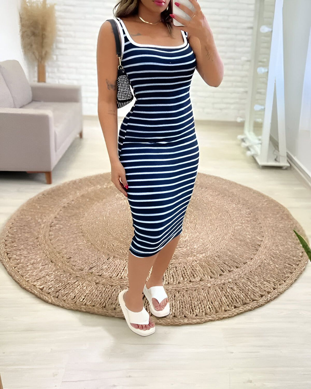 Une femme porte une robe mi-longue à fines rayures verticales noires et blanches. Elle porte également des sandales blanches à semelles compensées et un sac à main noir et blanc.