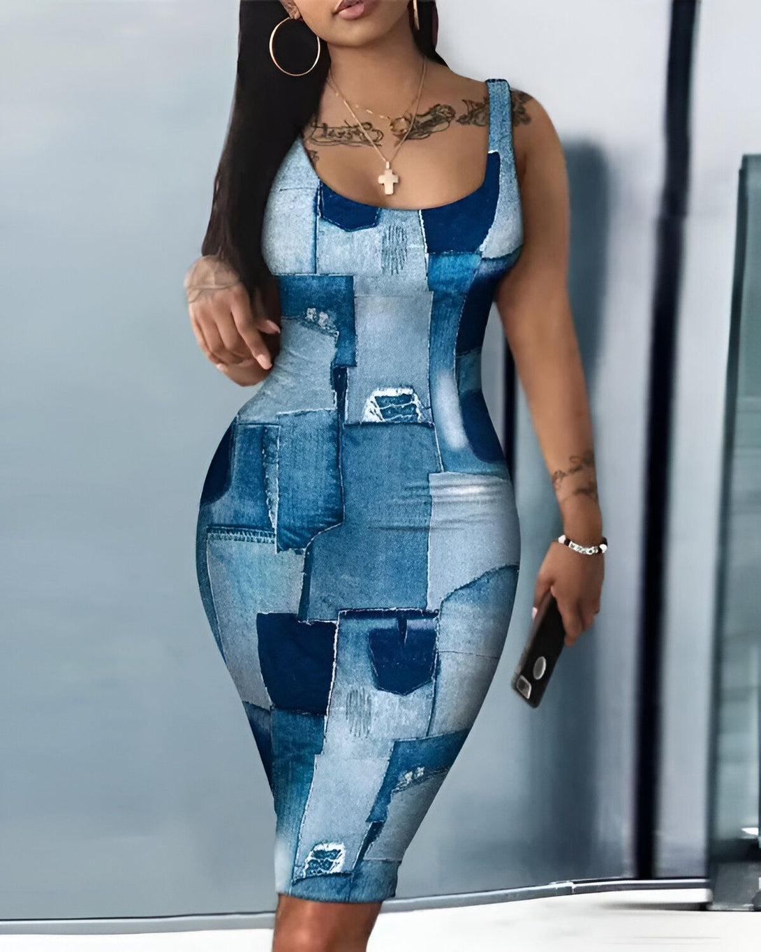 Une femme porte une robe moulante en jean bleu. La robe est courte et sans manches. Elle a un décolleté carré et des bretelles fines. La femme est debout, de face, et elle regarde l'appareil photo. Elle a les cheveux longs et noirs, et elle porte du maquillage.