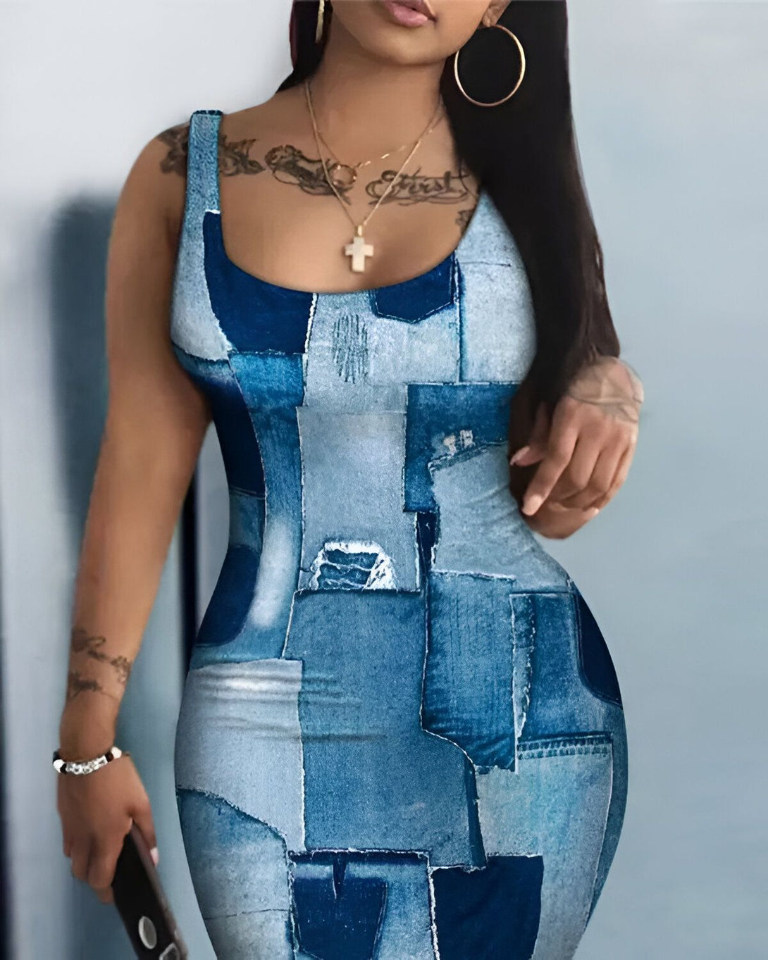 Une femme porte une robe moulante en jean bleu. La robe est courte et sans manches. Elle a un décolleté carré. La femme est debout, elle a les cheveux longs et noirs. Elle porte un collier avec une croix.