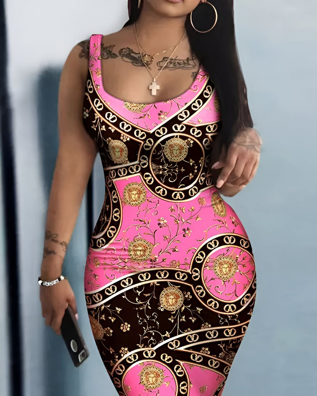 Une femme porte une robe rose et noire moulante. La robe est imprimée d'un motif de chaînes et de médaillons dorés. La femme est debout, de face, et tient un téléphone portable à la main.