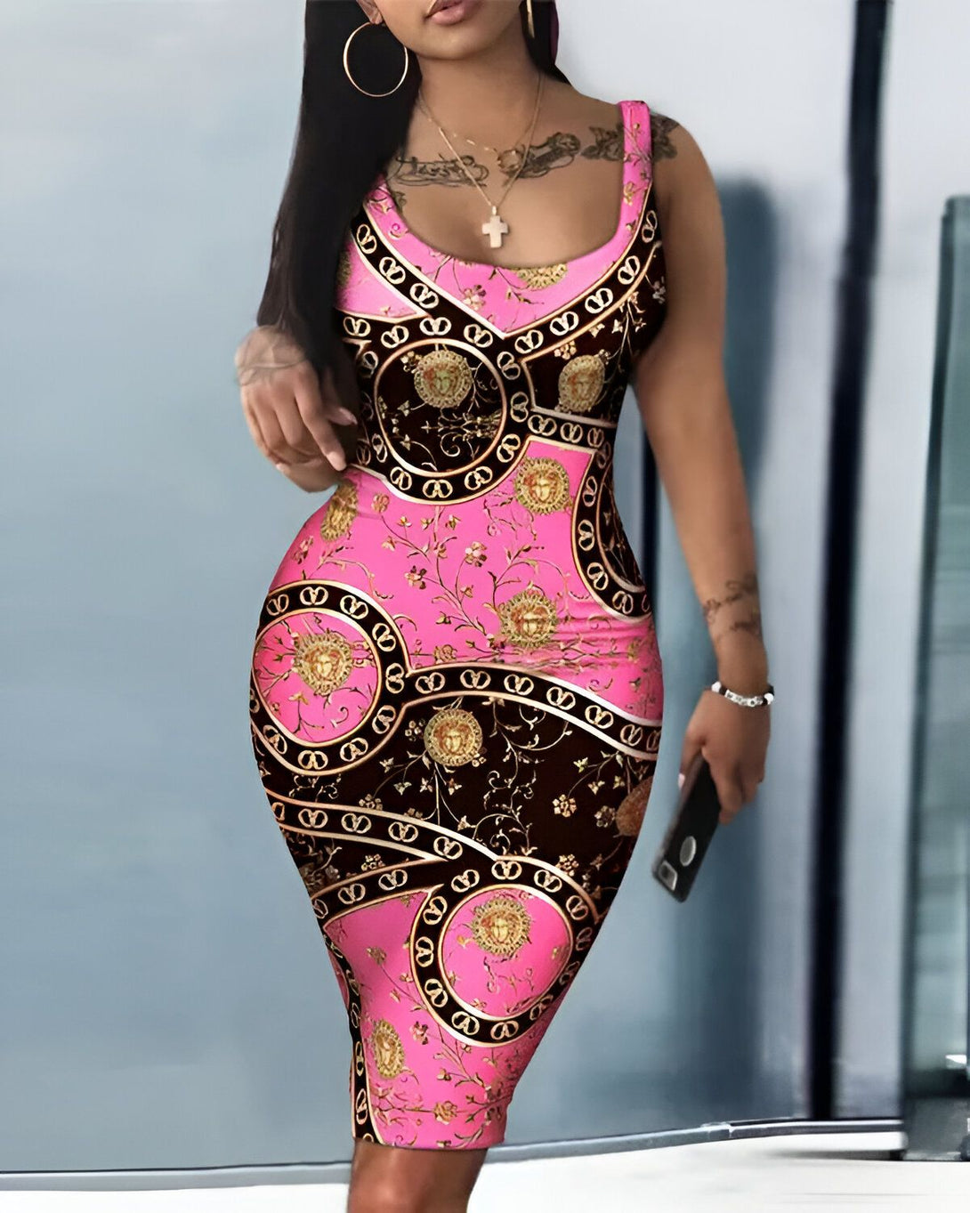 Une femme porte une robe rose et noire moulante. La robe est imprimée d'un motif de chaînes et de médaillons dorés. La femme est debout, de face, et elle regarde l'appareil photo. Elle a les cheveux longs et noirs, et elle porte un collier avec une croix.