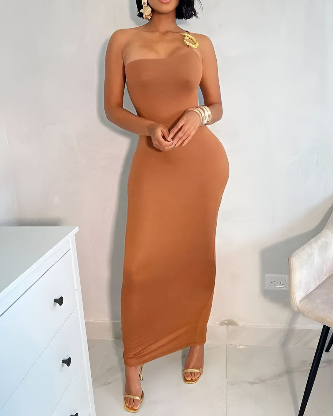 Une femme porte une robe moulante marron. La robe est sans bretelles et a une fente sur le côté. La femme est debout dans une pièce blanche avec un mur blanc en arrière-plan.