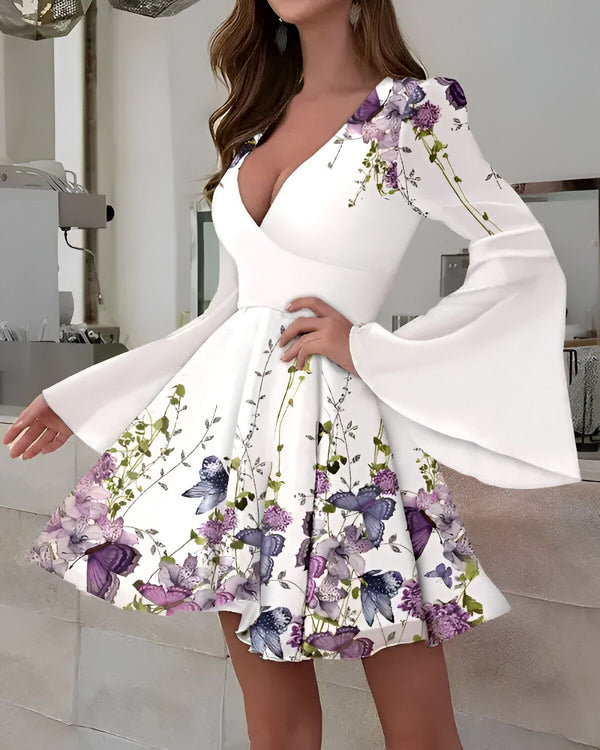 Une jeune femme porte une robe blanche à fleurs violettes et papillons. La robe est courte et évasée, avec un décolleté en V et des manches longues. La femme est debout, les cheveux longs et noirs.
