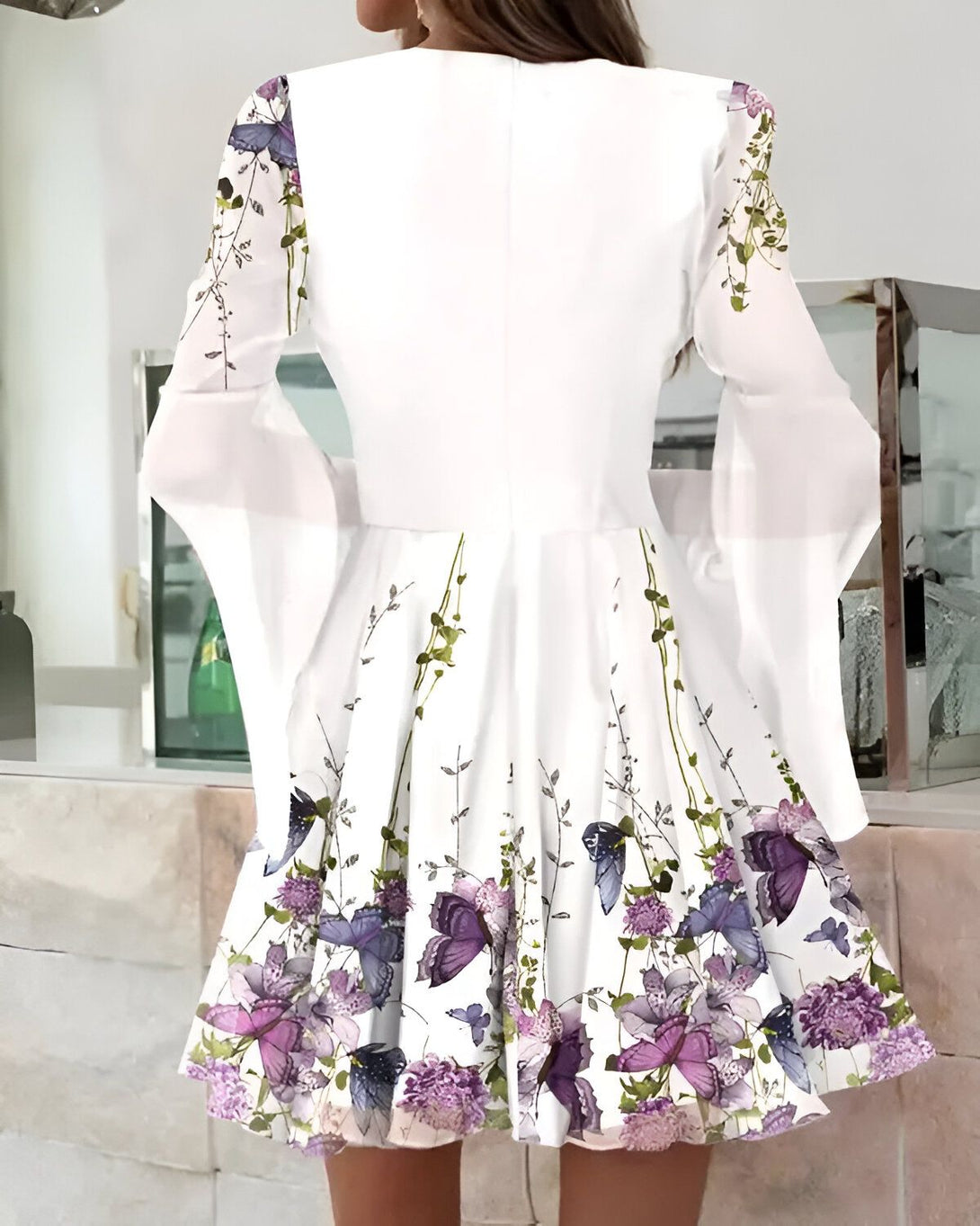 Une femme porte une robe blanche à motifs floraux et papillons. La robe est courte et évasée, avec des manches longues et évasées. Le dos de la robe est ouvert et la femme porte ses cheveux attachés en chignon.