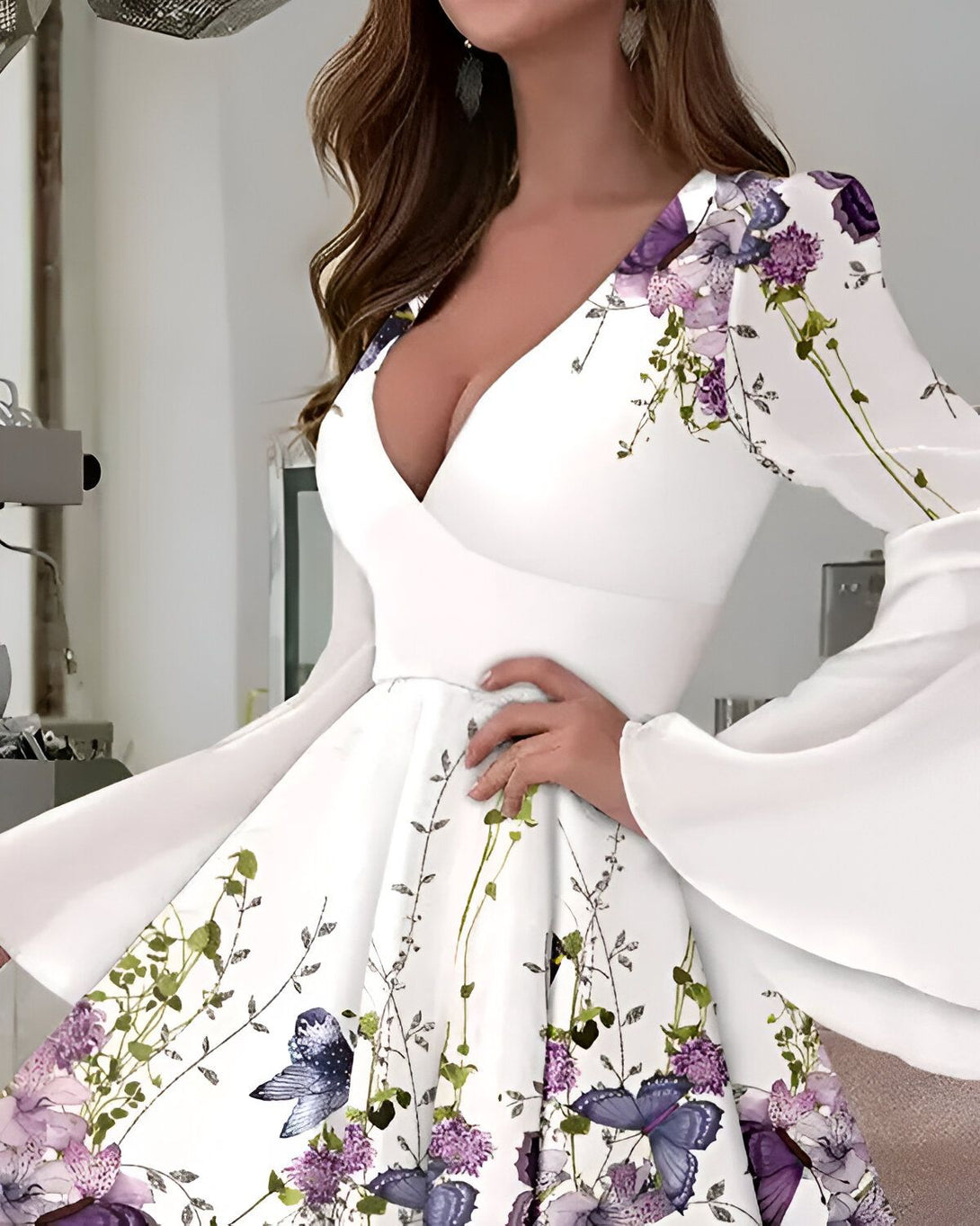 Une jeune femme porte une robe blanche à fleurs violettes et papillons. La robe est décolletée et a des manches longues. La femme est debout et a une main sur la hanche.