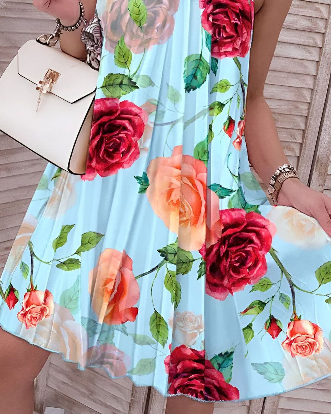 Une femme porte une robe plissée bleu clair à motif de roses rouges et de feuilles vertes. Elle porte également un sac à main blanc.