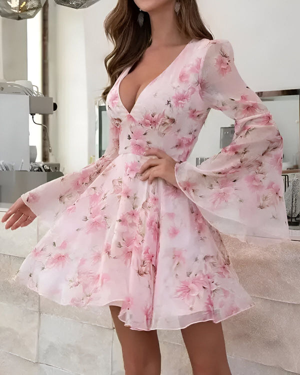 Une jeune femme porte une robe rose à fleurs. La robe est courte et évasée, avec un décolleté en V plongeant. Les manches sont longues et évasées. La femme est debout dans une cuisine, et elle a l'air heureuse et confiante.