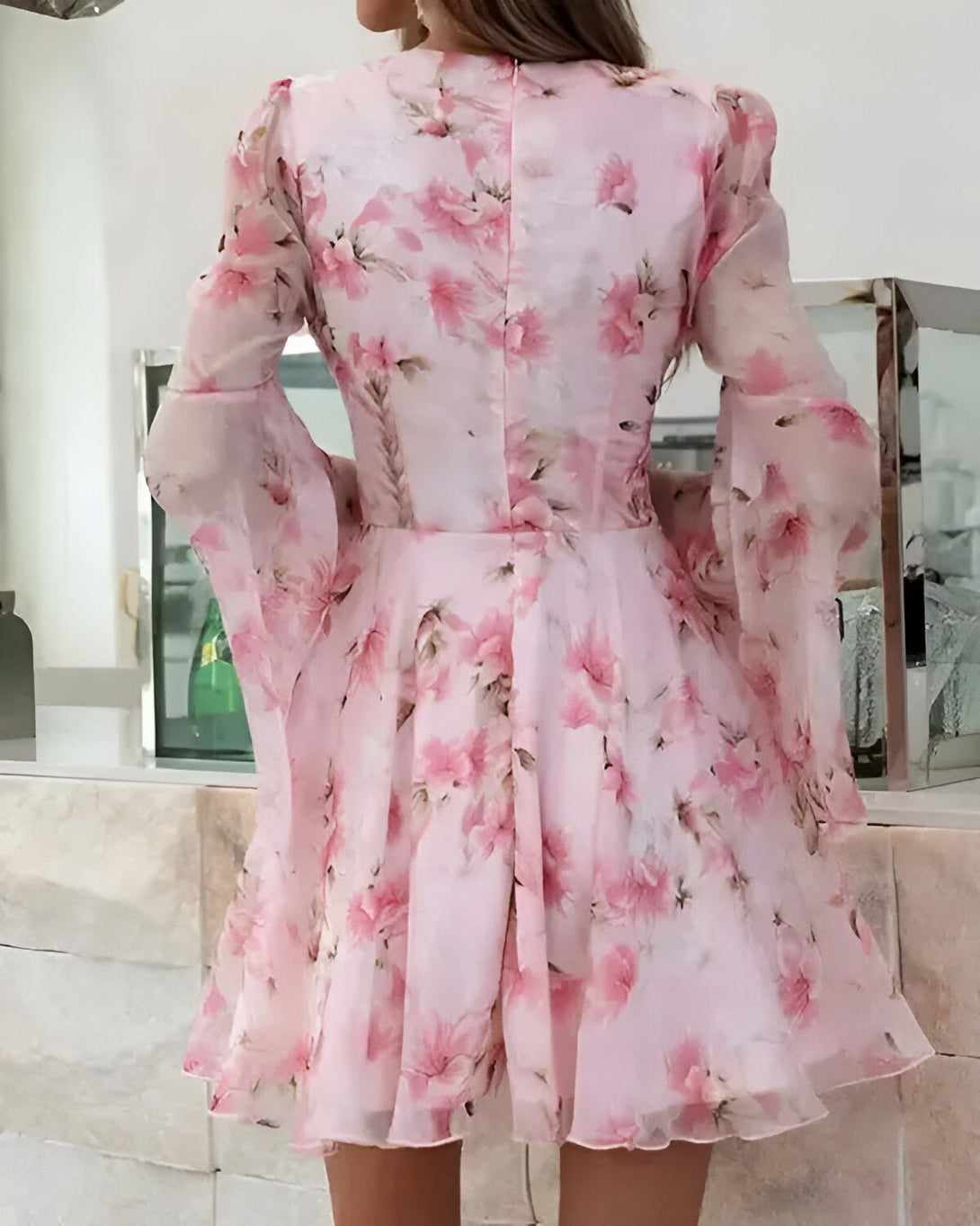 Une femme porte une robe rose à fleurs. La robe est courte et évasée, avec des manches longues et évasées. Le tissu est léger et transparent. La femme est vue de dos.