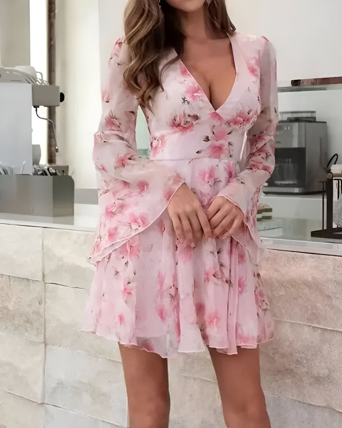 Une jeune femme portant une robe rose à fleurs. La robe est courte et a des manches longues. La femme est debout dans une cuisine et sourit à la caméra.
