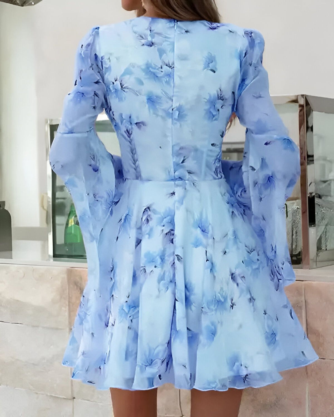 Une femme porte une robe bleue à fleurs. La robe est courte et évasée, avec des manches longues et évasées. Le tissu est léger et transparent. La femme est vue de dos.