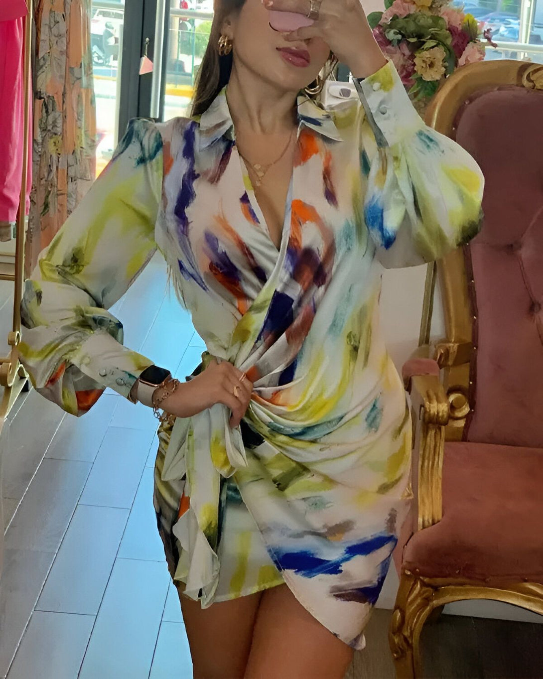 Une femme porte une robe portefeuille en soie imprimée. La robe est multicolore et présente un motif abstrait. La femme est debout dans un magasin et se regarde dans un miroir.