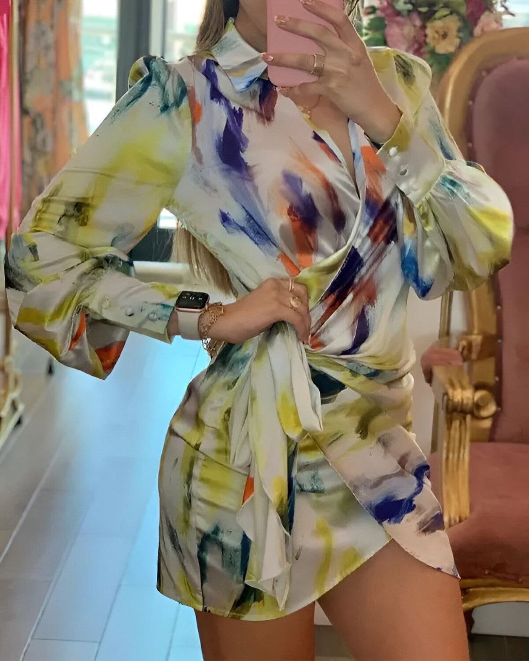 Une femme porte une robe portefeuille en satin imprimée de motifs abstraits. La robe est nouée sur le côté et possède un col montant. La femme est debout dans un magasin et se regarde dans le miroir.