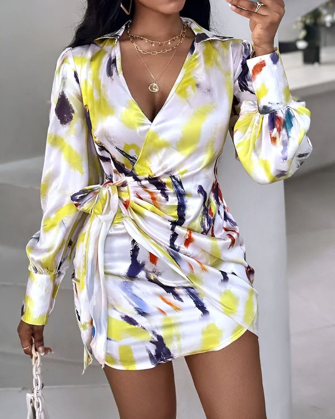 Une femme porte une robe portefeuille en satin imprimée de motifs abstraits. La robe est jaune et blanche, avec des touches de bleu, de vert et de rose. La femme est debout, la main sur la hanche, et regarde l'appareil photo. Elle porte des talons hauts blancs et un collier doré.