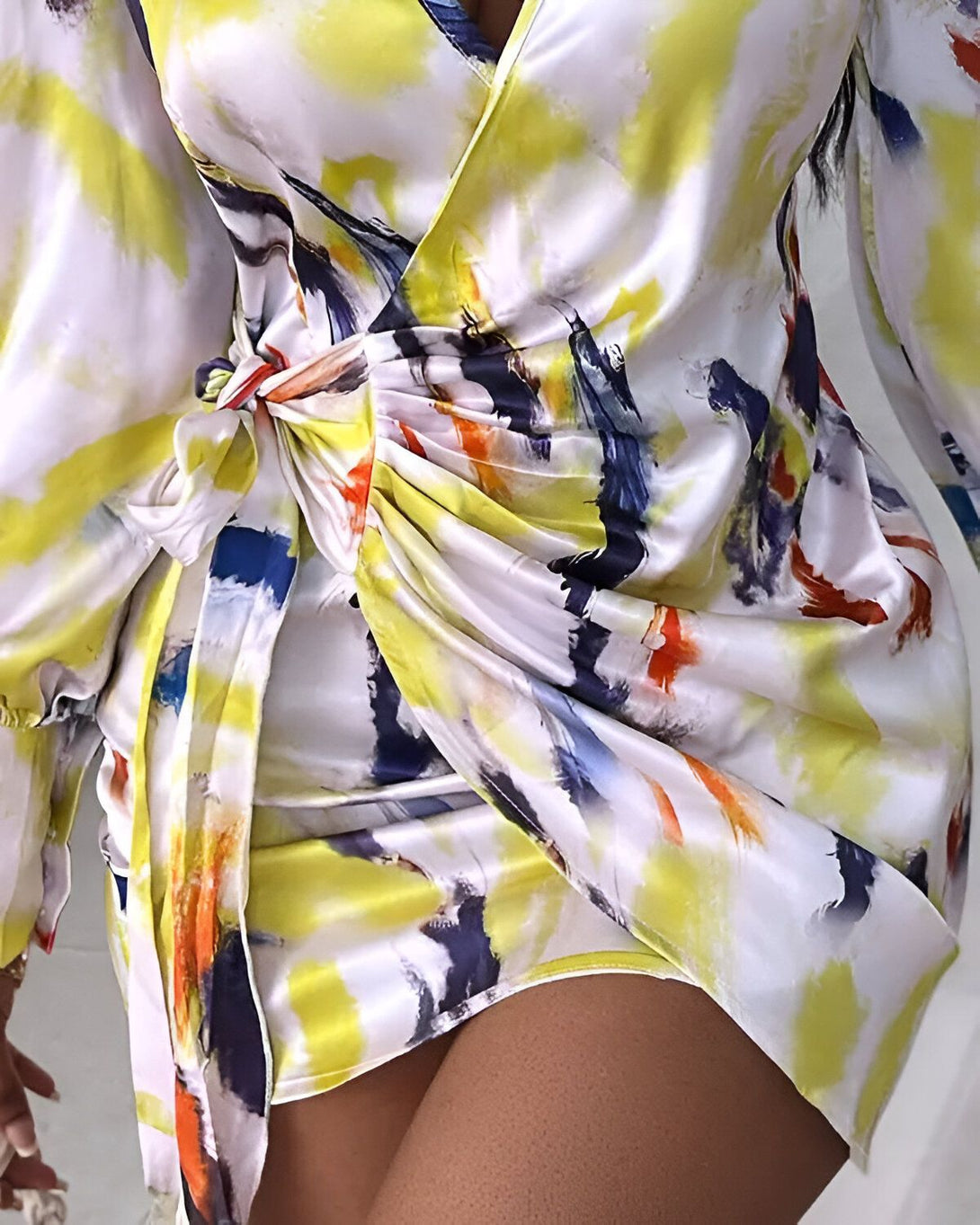 Une femme porte une robe portefeuille en satin imprimée de motifs abstraits. La robe est jaune et blanche, avec des touches de bleu, de vert et de rose. La femme est vue de dos, et ses cheveux sont lâchés.