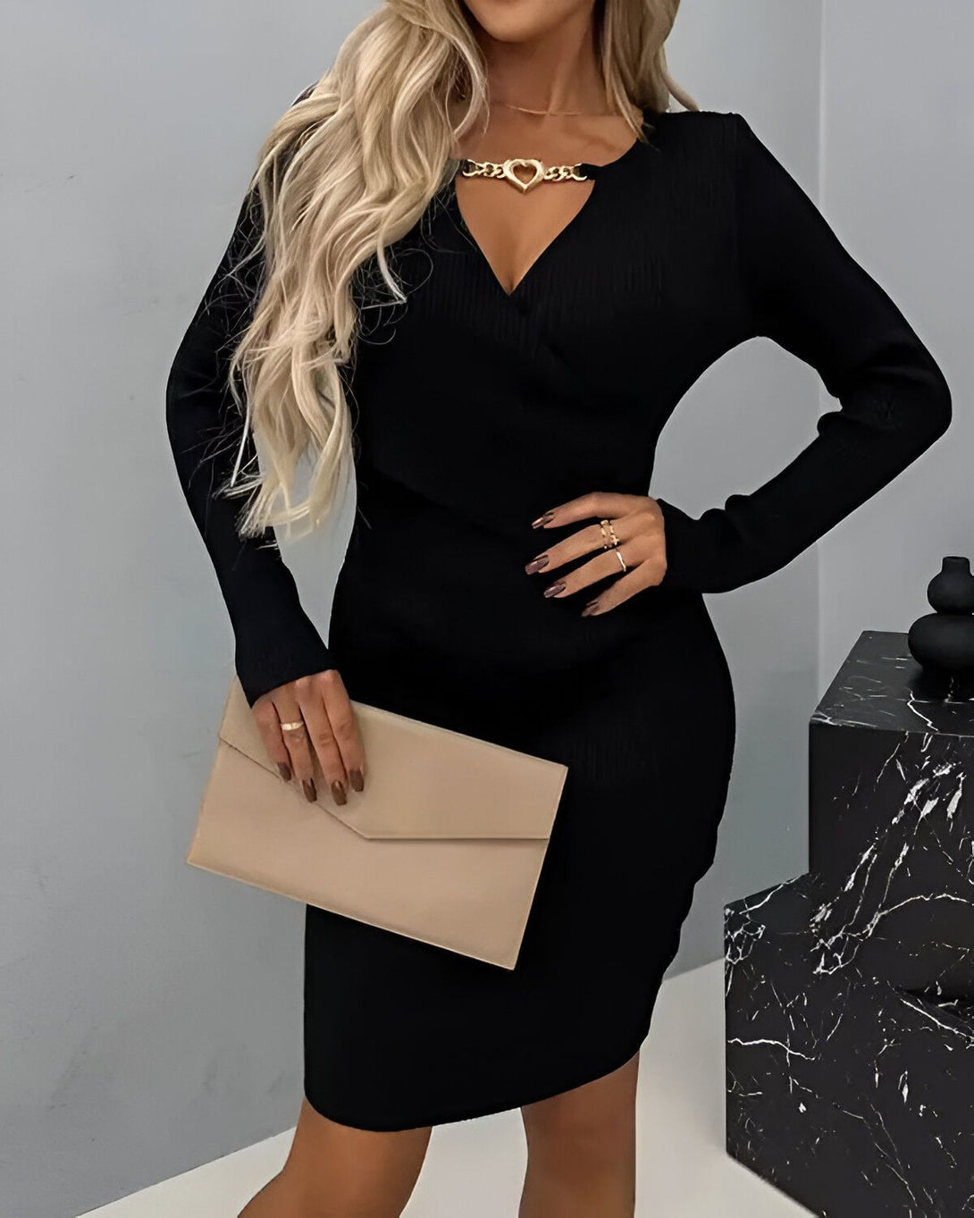 Une femme porte une robe noire moulante à manches longues avec un décolleté en V et une chaîne dorée en forme de cœur. Elle porte également un sac à main beige et des talons noirs.