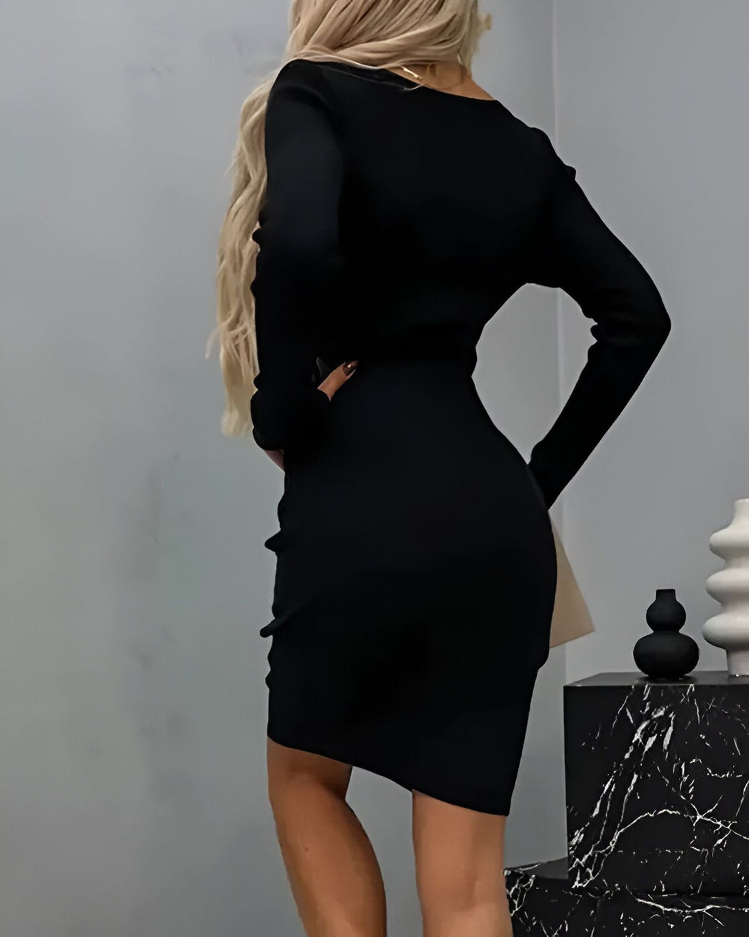 Une femme porte une robe noire moulante à manches longues. La robe est courte et met en valeur ses courbes. Elle a les cheveux blonds et porte des talons noirs. Elle est debout dans une pièce avec un mur gris et un sol en marbre.