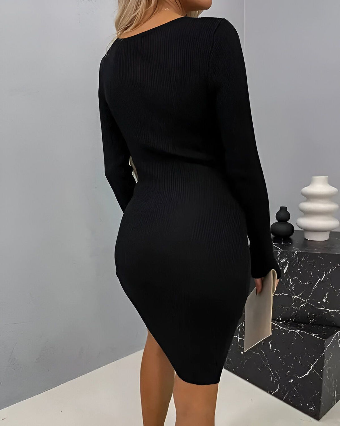 Une femme porte une robe noire moulante à manches longues. La robe est fendue sur le côté. La femme est blonde et a les cheveux longs. Elle porte des talons noirs.