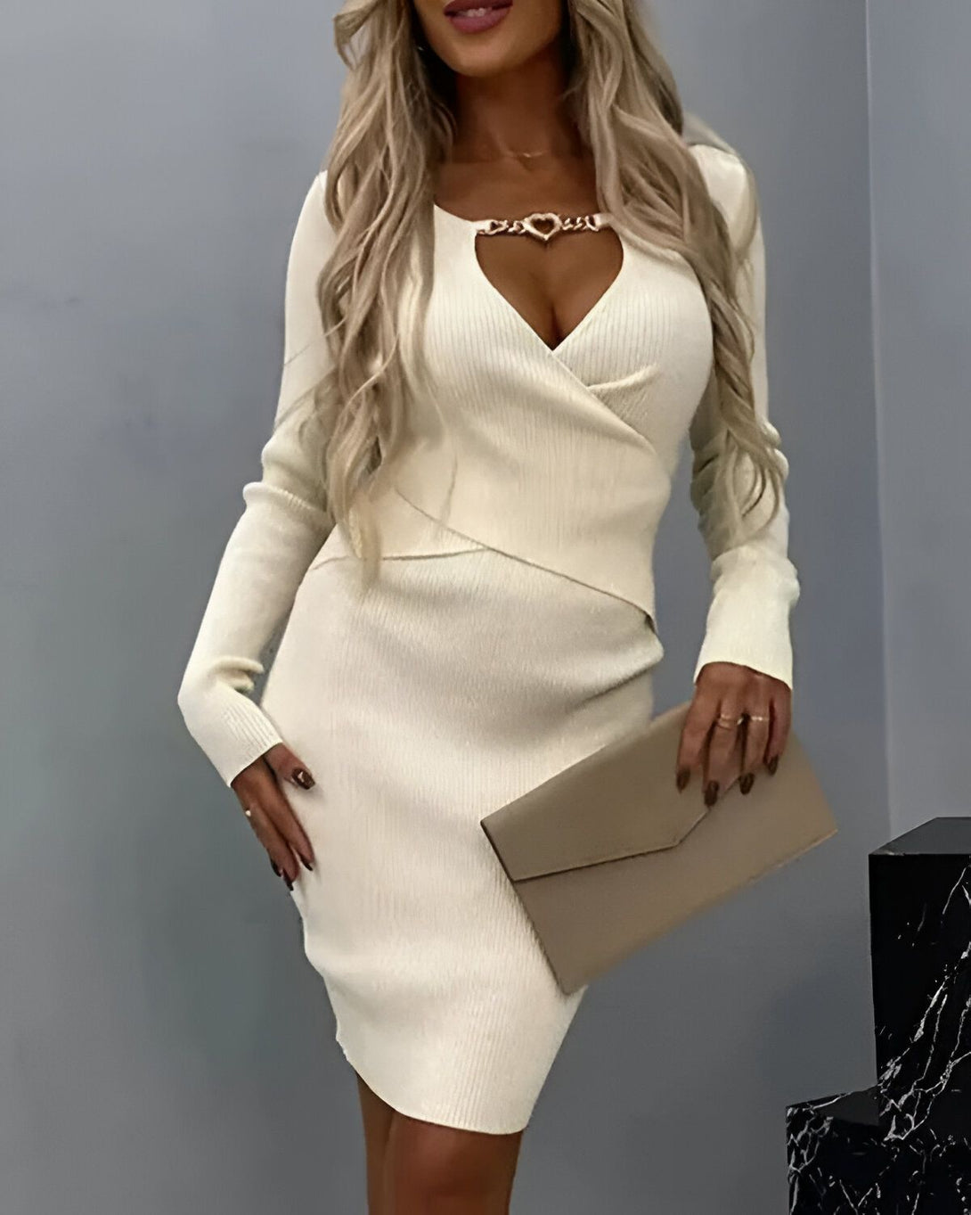 Une femme porte une robe blanche moulante avec un décolleté en V et une chaîne dorée en forme de cœur. Elle porte également un sac à main beige.