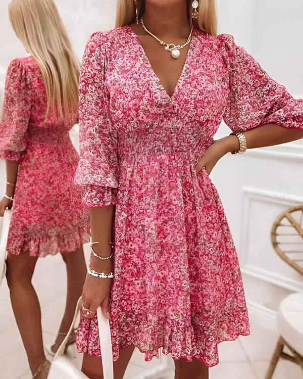 Une jeune femme porte une robe rose à fleurs. La robe est courte et a un décolleté en V. Les manches sont bouffantes et la jupe est évasée. La femme porte également un collier et des bracelets.