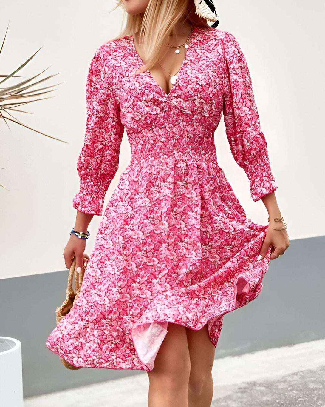 Une jeune femme porte une robe rose à fleurs. La robe est courte et évasée, avec des manches longues et un col en V. La femme est blonde et porte un chapeau de paille. Elle est souriante et marche dans la rue.
