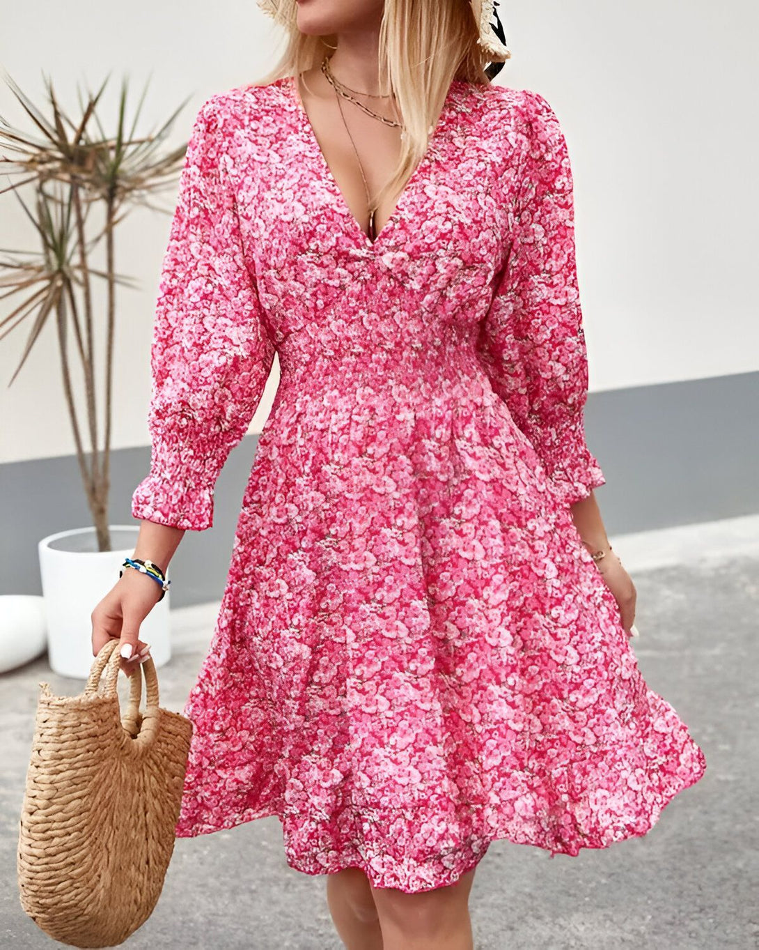 Une femme porte une robe rose à fleurs. La robe est courte et a un décolleté en V. Les manches sont bouffantes et la jupe est évasée. La femme porte un chapeau de paille et des sandales. Elle porte également un sac à main en osier.